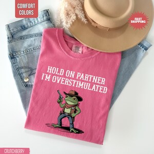 May include: A pink t-shirt with the text "HOLD ON PARTNER I'M OVERSTIMULATED" and a cartoon frog wearing a cowboy hat and holding a gun. The shirt is folded on top of blue jeans and a tan hat.