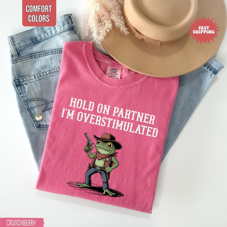 May include: A pink t-shirt with the text "HOLD ON PARTNER I'M OVERSTIMULATED" and a cartoon frog wearing a cowboy hat and holding a gun. The shirt is folded on top of blue jeans and a tan hat.