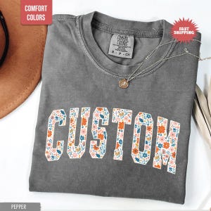 K&ouml;nnte beinhalten: Ein graues Comfort Colors T-Shirt mit dem Wort "CUSTOM" in einem floralen Design. Das Shirt hat einen Rundhalsausschnitt und kurze &Auml;rmel. Eine silberne Halskette mit Anh&auml;nger liegt auf dem Shirt.