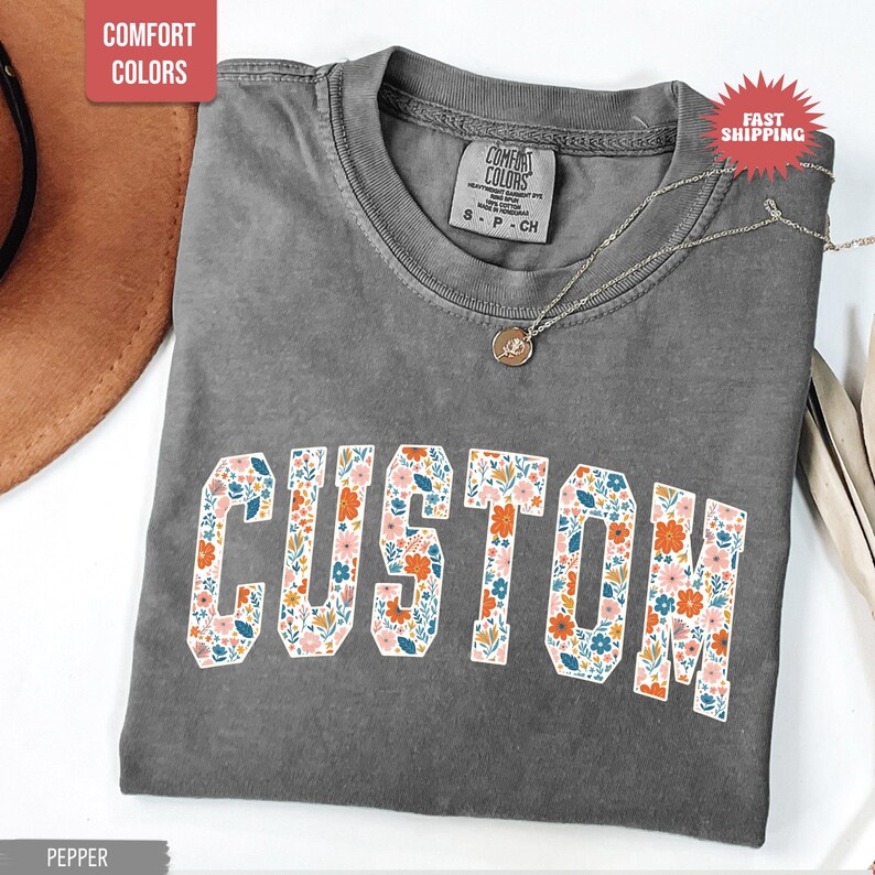 K&ouml;nnte beinhalten: Ein graues Comfort Colors T-Shirt mit dem Wort "CUSTOM" in einem floralen Design. Das Shirt hat einen Rundhalsausschnitt und kurze &Auml;rmel. Eine silberne Halskette mit Anh&auml;nger liegt auf dem Shirt.
