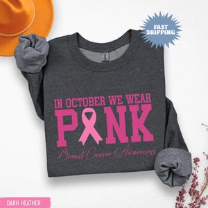 May include: Dark heather gray sweatshirt with the text "IN OCTOBER WE WEAR PINK" in pink block letters. A pink ribbon graphic is centered within the word "PINK". The words "Breast Cancer Awareness" are below the word "PINK".