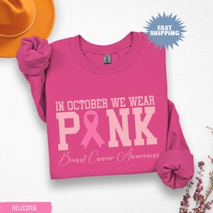 May include: A bright pink sweatshirt with the text "IN OCTOBER WE WEAR PINK" and "Breast Cancer Awareness" in white and light pink. A pink ribbon graphic is also featured. The sweatshirt has long sleeves.