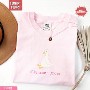 May include: A light pink Comfort Colors t-shirt with a cartoon goose illustration and the text "silly mama goose" in pink. A gold necklace with a pendant is draped on the shirt. The shirt is folded on a white surface.
