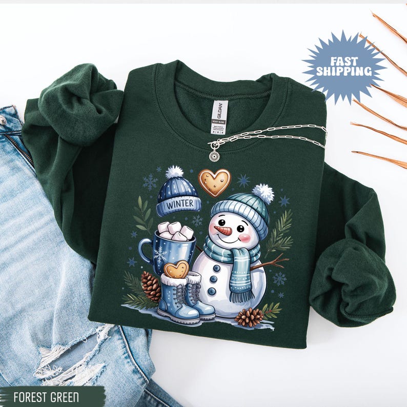 May include: Forest green sweatshirt featuring a whimsical snowman design with a blue hat, scarf, and boots. The graphic includes a mug of marshmallows, gingerbread cookies, and the word "WINTER". A silver necklace is draped on the sweatshirt.
