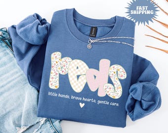 Puede incluir: Sudadera azul índigo con la palabra "reds" en estampados florales y cuadros vichy. El texto "little hands, brave hearts, gentle care" está debajo. También se ven un collar plateado y un gráfico de "Fast Shipping".