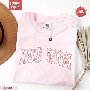 May include: A light pink t-shirt with the words "DOG MOM" in a floral pattern. The shirt has a crew neck and short sleeves. A gold necklace with a pendant is draped on the shirt. The words "COMFORT COLORS" are printed on the shirt's tag.