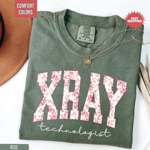 May include: Moss green t-shirt with the word "XRAY" in a floral design and the word "technologist" in white script. The shirt has a crew neck and a small tag that says "Comfort Colors". A silver necklace with a pendant is on the shirt.