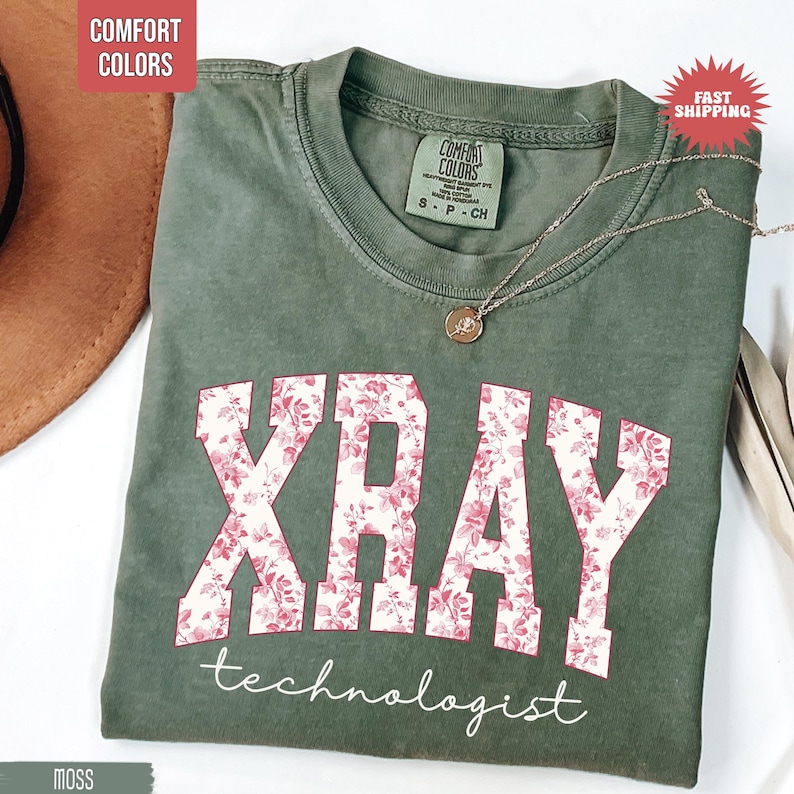 May include: Moss green t-shirt with the word "XRAY" in a floral design and the word "technologist" in white script. The shirt has a crew neck and a small tag that says "Comfort Colors". A silver necklace with a pendant is on the shirt.