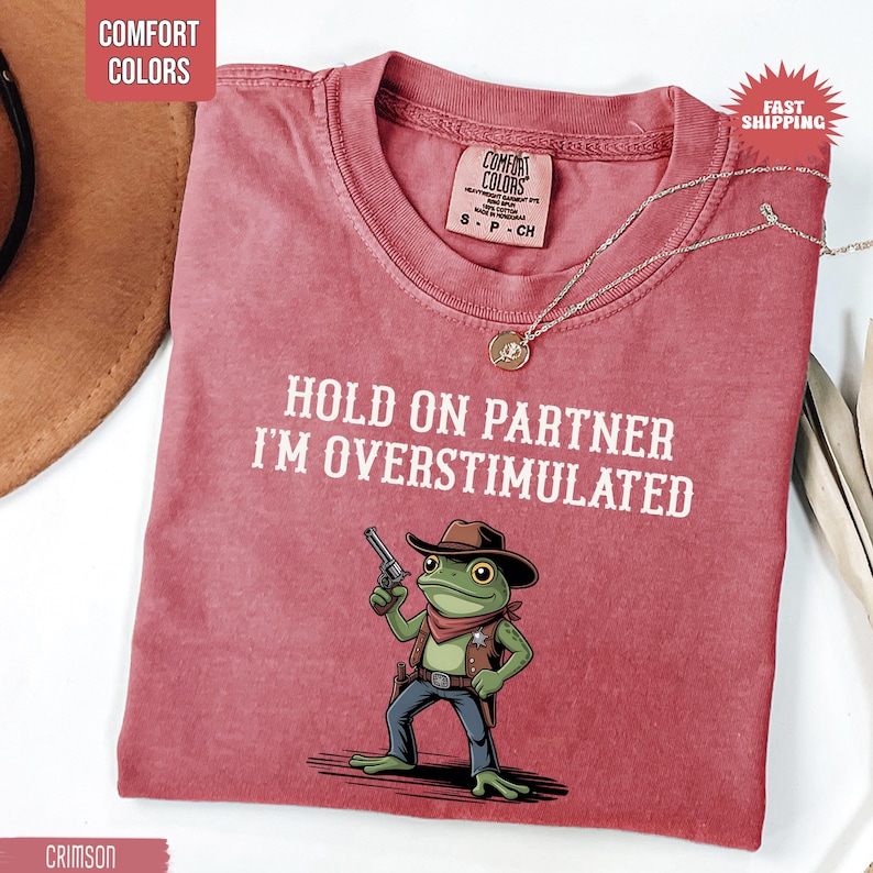 May include: Crimson Comfort Colors t-shirt with the text "HOLD ON PARTNER I'M OVERSTIMULATED" and a cartoon frog wearing a cowboy hat and holding a gun. A gold necklace is draped across the shirt.