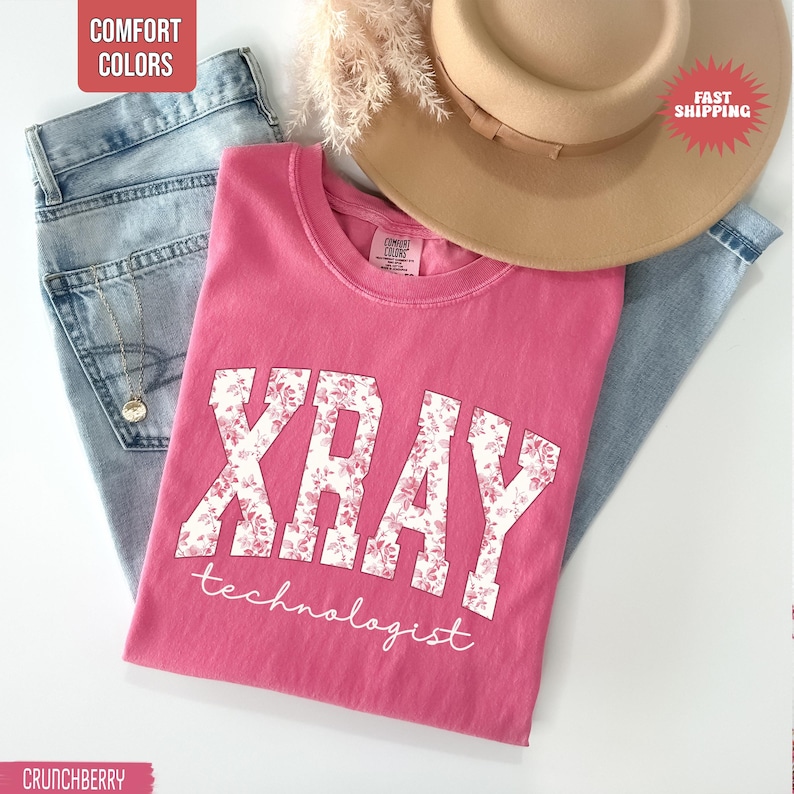 May include: A pink t-shirt with the words "XRAY technologist" in white floral lettering. The shirt is paired with light blue jeans and a tan hat. The shirt is labeled "Comfort Colors".