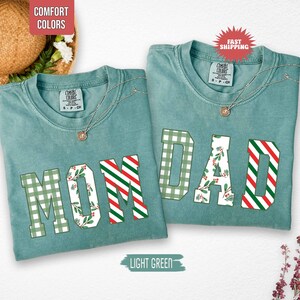 May include: Two light green t-shirts with the words "MOM" and "DAD" in a mix of green plaid, red and white stripes, and floral patterns. Each shirt has a silver necklace. The shirts are labeled "Comfort Colors".