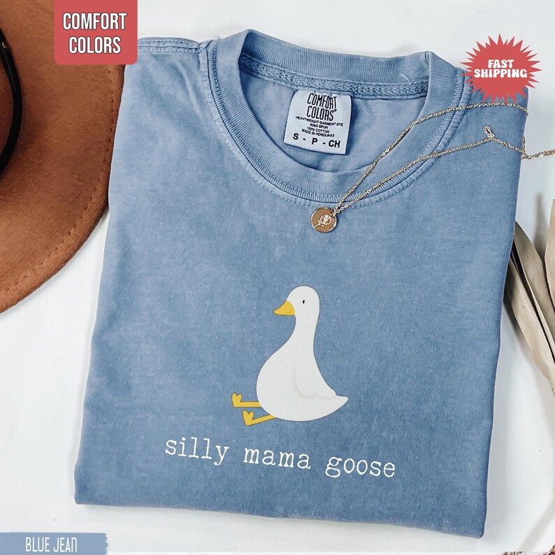 May include: Blue jean Comfort Colors t-shirt with a white goose graphic and the words "silly mama goose" in white. A gold necklace with a pendant rests on the shirt. The shirt is a size small.
