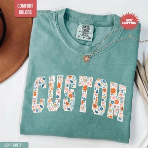 K&ouml;nnte beinhalten: Hellgr&uuml;nes T-Shirt mit dem Wort "CUSTOM" in einem floralen Design. Das Shirt hat einen Rundhalsausschnitt und kurze &Auml;rmel. Eine silberne Halskette mit Anh&auml;nger ist &uuml;ber dem Shirt drapiert. Das Shirt ist mit "Comfort Colors" gekennzeichnet.