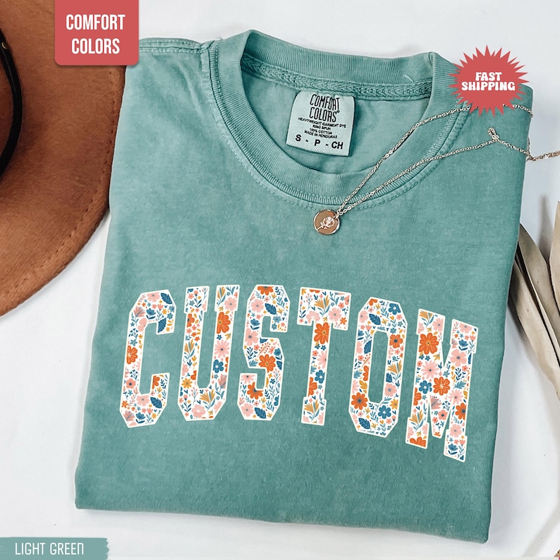 K&ouml;nnte beinhalten: Hellgr&uuml;nes T-Shirt mit dem Wort "CUSTOM" in einem floralen Design. Das Shirt hat einen Rundhalsausschnitt und kurze &Auml;rmel. Eine silberne Halskette mit Anh&auml;nger ist &uuml;ber dem Shirt drapiert. Das Shirt ist mit "Comfort Colors" gekennzeichnet.