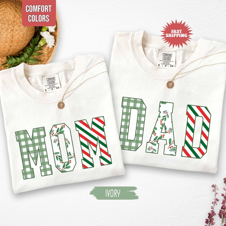 May include: Two ivory-colored t-shirts with the words "MOM" and "DAD" in green, red, and white lettering. The "MOM" shirt has a green plaid "M" and "O", with a floral design in the "O". The "DAD" shirt has a green plaid "D" and a floral design in the "A".