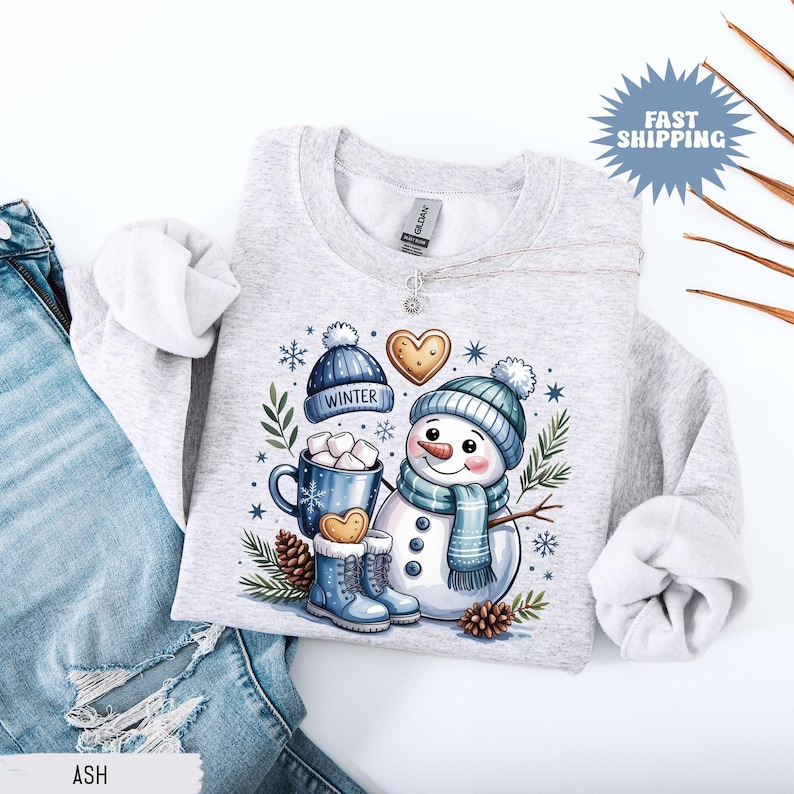 May include: Light gray sweatshirt featuring a whimsical snowman design. The illustration includes a snowman wearing a blue hat and scarf, a mug of hot chocolate, and the word "WINTER" on a hat. The design is surrounded by snowflakes and pinecones.