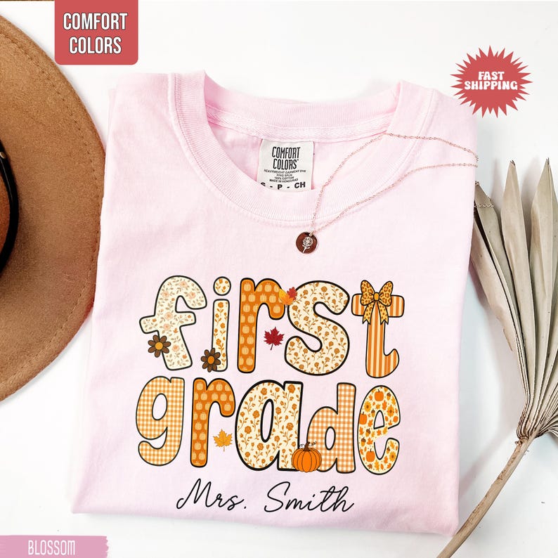 May include: A light pink t-shirt with the words "first grade" in a fall-themed design. The design includes floral patterns, pumpkins, and leaves. The name "Mrs. Smith" is printed below the words. A gold necklace is draped over the shirt.