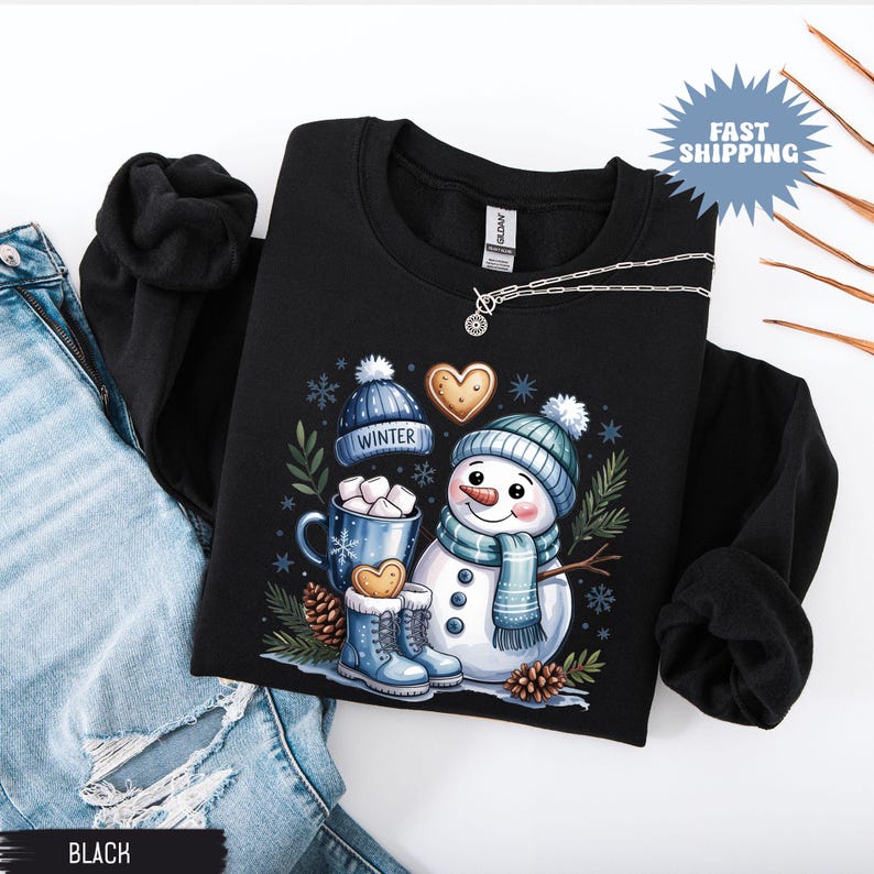 May include: Black sweatshirt featuring a whimsical snowman design. The snowman wears a blue hat and scarf, with a mug of hot cocoa and gingerbread cookies. The word "WINTER" is on a blue hat. A silver necklace is also visible.