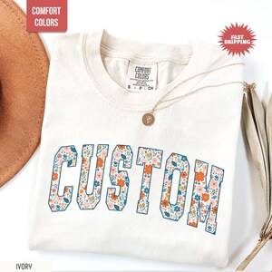 K&ouml;nnte beinhalten: Elfenbeinfarbenes T-Shirt mit dem Wort "CUSTOM" in einem floralen Design. Das Shirt hat einen Rundhalsausschnitt und kurze &Auml;rmel. Eine goldene Halskette mit Anh&auml;nger liegt auf dem Shirt. Die W&ouml;rter "COMFORT COLORS" und "FAST SHIPPING" sind ebenfalls sichtbar.