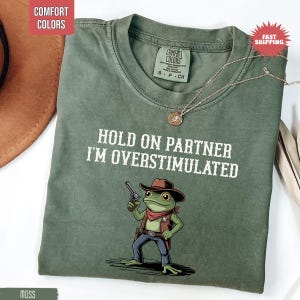 May include: A moss green t-shirt with the text "HOLD ON PARTNER I'M OVERSTIMULATED" and a cartoon frog wearing a cowboy hat and holding a gun. The shirt has a crew neck and short sleeves. A silver necklace with a pendant is also visible.
