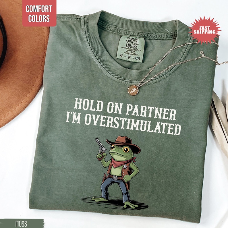 May include: A moss green t-shirt with the text "HOLD ON PARTNER I'M OVERSTIMULATED" and a cartoon frog wearing a cowboy hat and holding a gun. The shirt has a crew neck and short sleeves. A silver necklace with a pendant is also visible.