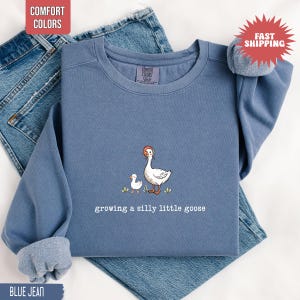 May include: Blue jean colored sweatshirt with a goose and gosling graphic. The text "growing a silly little goose" is below the illustration. The sweatshirt has a crew neck and long sleeves. The words "Comfort Colors" and "Fast Shipping" are in the image.