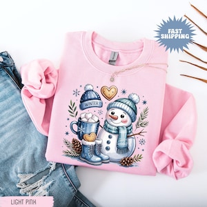 May include: Light pink sweatshirt featuring a snowman graphic with a blue hat, scarf, and boots. The design includes a mug of hot cocoa, a gingerbread heart, and the word "Winter" on a blue hat. The sweatshirt is displayed on a pair of blue jeans.