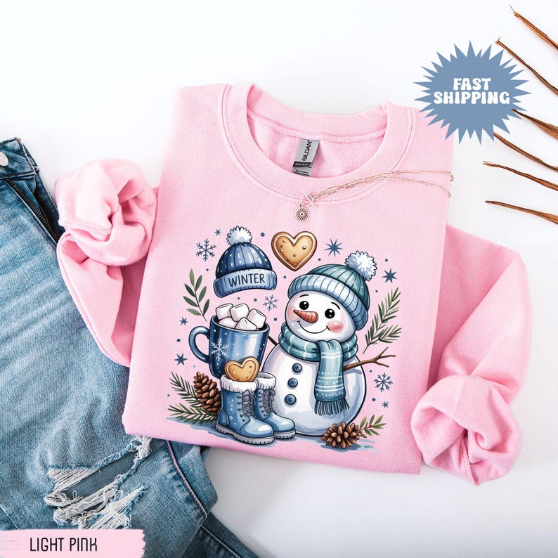 May include: Light pink sweatshirt featuring a snowman graphic with a blue hat, scarf, and boots. The design includes a mug of hot cocoa, a gingerbread heart, and the word "Winter" on a blue hat. The sweatshirt is displayed on a pair of blue jeans.