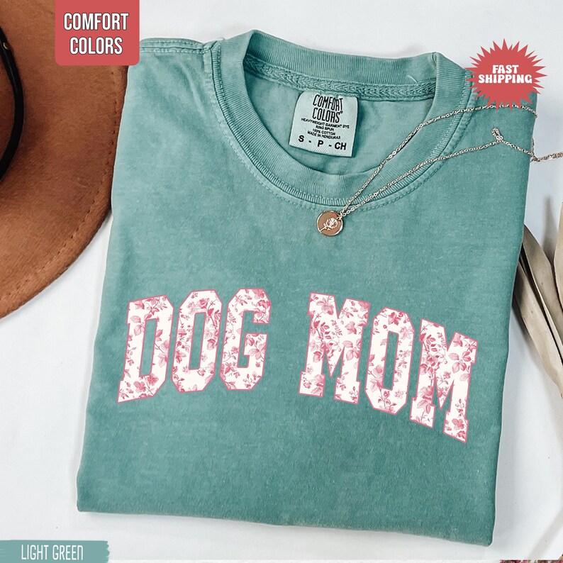 May include: Light green t-shirt with the text "DOG MOM" in a floral pattern. The shirt has a crew neck and a small tag on the collar. A silver necklace with a pendant is draped on the shirt.