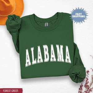 May include: Forest green sweatshirt with the word "ALABAMA" in white, distressed, block letters. The sweatshirt has long sleeves and a crew neck. A Fast Shipping graphic is in the upper right corner.