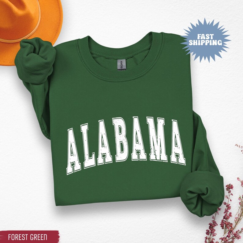 May include: Forest green sweatshirt with the word "ALABAMA" in white, distressed, block letters. The sweatshirt has long sleeves and a crew neck. A Fast Shipping graphic is in the upper right corner.