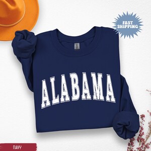 May include: Navy blue sweatshirt with the word "ALABAMA" in distressed white lettering. The sweatshirt is folded, with the sleeves bunched up. A gold-colored hat is visible in the upper left corner.