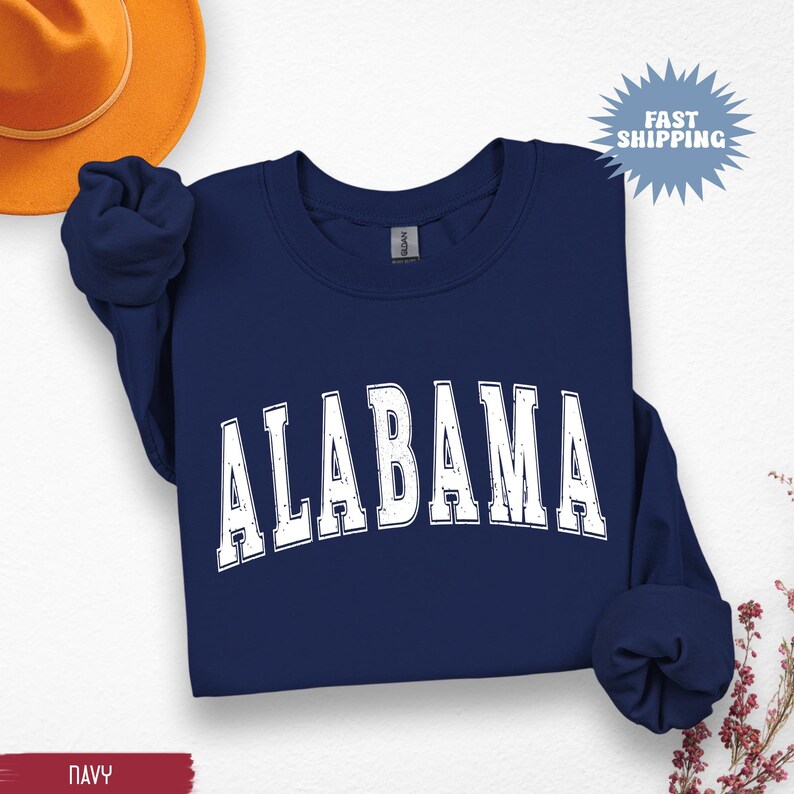 May include: Navy blue sweatshirt with the word "ALABAMA" in distressed white lettering. The sweatshirt is folded, with the sleeves bunched up. A gold-colored hat is visible in the upper left corner.