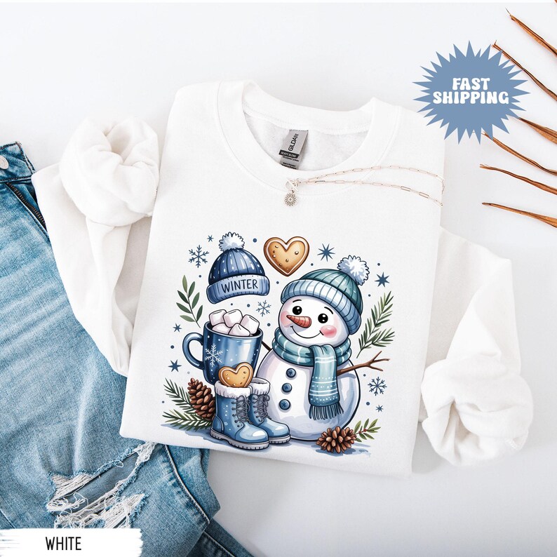 May include: White sweatshirt featuring a whimsical winter design. The graphic includes a snowman with a blue hat and scarf, a mug of marshmallows, and winter boots. A blue hat with the word "WINTER" is also featured.