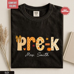 May include: Black t-shirt with the word "pre-k" in orange and white fall-themed patterns, including pumpkins and leaves. The name "Mrs. Smith" is written below the text. The shirt has a round neckline.