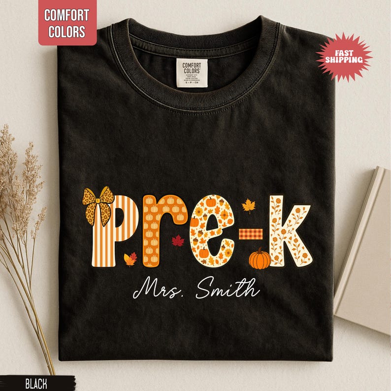 May include: Black t-shirt with the word "pre-k" in orange and white fall-themed patterns, including pumpkins and leaves. The name "Mrs. Smith" is written below the text. The shirt has a round neckline.