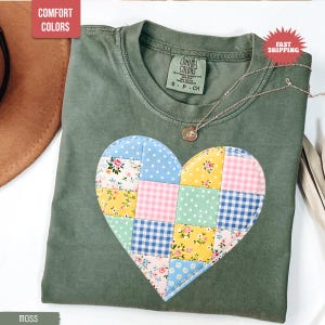 Puede incluir: Camiseta Comfort Colors verde musgo con un diseño de corazón de patchwork. El corazón está hecho de varias telas estampadas en azul, rosa, amarillo y blanco. También se ve un collar plateado con un pequeño colgante.
