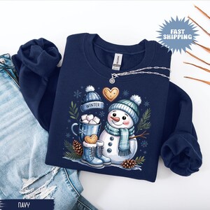 May include: Navy blue sweatshirt featuring a snowman design with a blue hat and scarf, holding a mug of hot chocolate. The design includes the word "WINTER" on a hat, gingerbread cookies, and snowflakes. A silver necklace is draped across the sweatshirt.