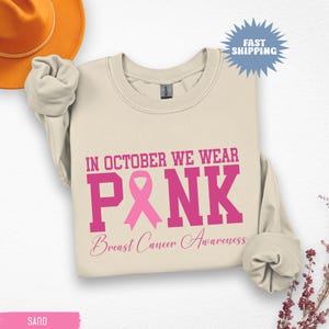 May include: Sand-colored sweatshirt with the text "IN OCTOBER WE WEAR PINK" in pink block letters, with a pink ribbon graphic. Below, the words "Breast Cancer Awareness" are written in a cursive font. A Fast Shipping sticker is in the upper right.