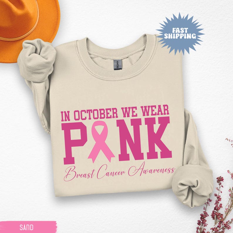 May include: Sand-colored sweatshirt with the text "IN OCTOBER WE WEAR PINK" in pink block letters, with a pink ribbon graphic. Below, the words "Breast Cancer Awareness" are written in a cursive font. A Fast Shipping sticker is in the upper right.