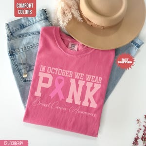 May include: A pink t-shirt featuring the text IN OCTOBER WE WEAR PINK with a pink ribbon replacing the letter I and the words Breast Cancer Awareness underneath. The shirt is displayed with light blue denim jeans and a tan wide-brimmed hat.