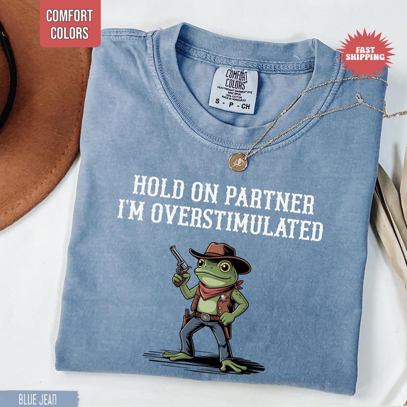 May include: Blue jean-colored Comfort Colors t-shirt with the text "HOLD ON PARTNER I'M OVERSTIMULATED" and a cartoon frog wearing a cowboy hat and holding a gun. A gold necklace is draped on the shirt.