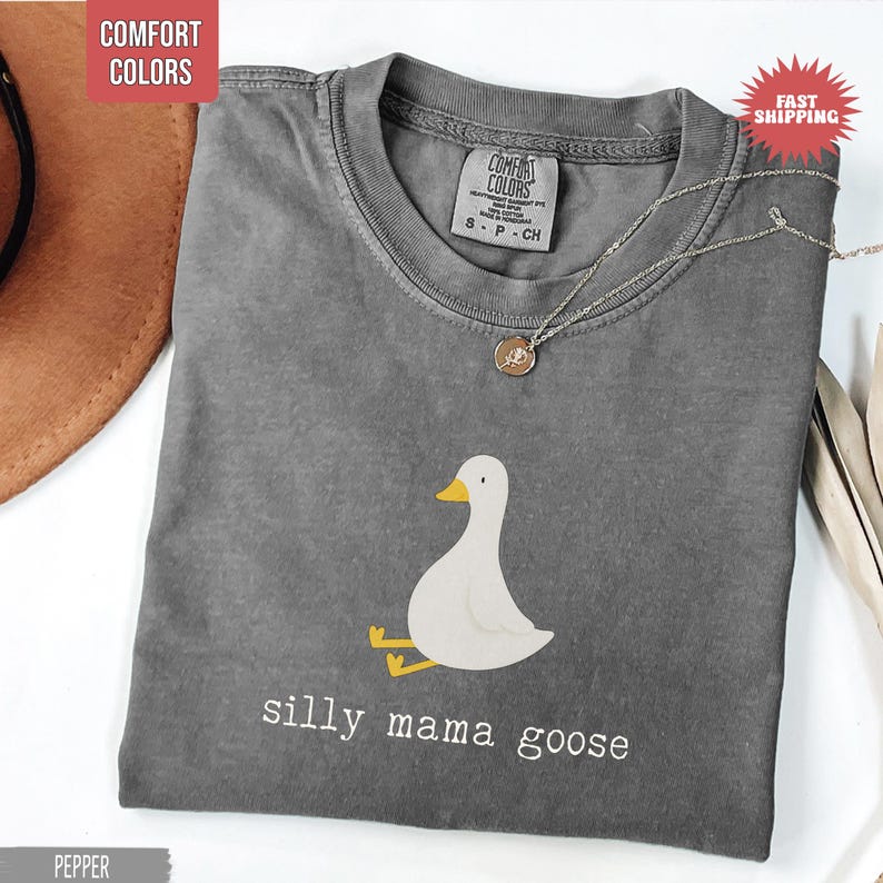 May include: A gray Comfort Colors t-shirt with a graphic of a white goose with yellow feet and beak. The text "silly mama goose" is printed below the goose. A silver necklace with a pendant is on the shirt.