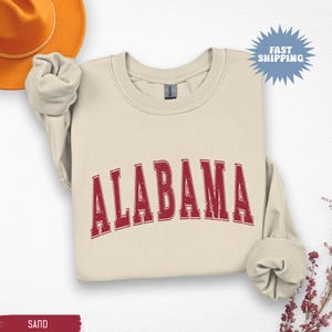 May include: Sand-colored sweatshirt with the word "ALABAMA" in a distressed, red, collegiate-style font. The sweatshirt has long sleeves and a crew neck. A brown hat is visible in the upper left corner. The words "FAST SHIPPING" are in a blue burst.