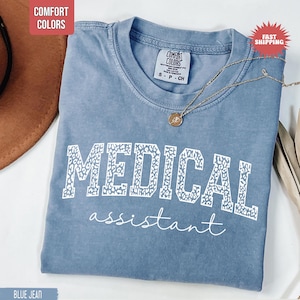 May include: Blue jean coloured t-shirt with the words "MEDICAL ASSISTANT" in white. The word "MEDICAL" has a leopard print design. A gold necklace with a pendant is on the shirt. The shirt has a Comfort Colors tag.