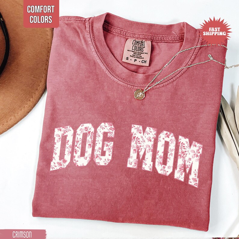 May include: Crimson-colored t-shirt with the text "DOG MOM" in white block letters filled with a floral pattern. The shirt has a crew neck and a small tag at the neckline. A silver necklace with a pendant is draped across the shirt.