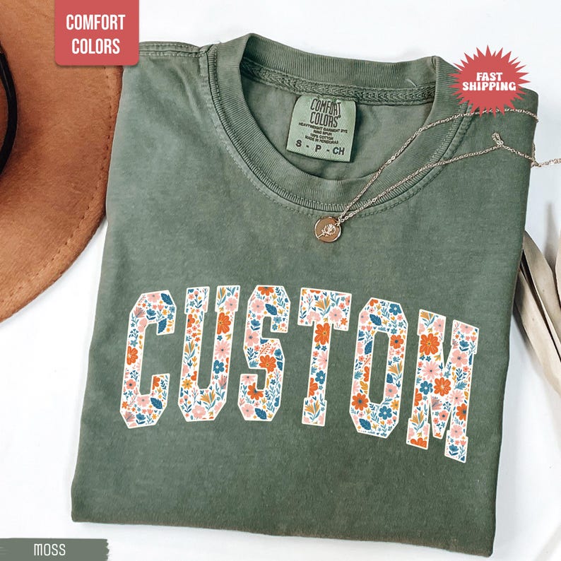 K&ouml;nnte beinhalten: Moosgr&uuml;nes T-Shirt mit dem Wort "CUSTOM" in einem floralen Design. Das Shirt hat einen Rundhalsausschnitt und kurze &Auml;rmel. Eine silberne Halskette mit Anh&auml;nger ist &uuml;ber dem Shirt drapiert. Das Shirt ist mit "Comfort Colors" gekennzeichnet.