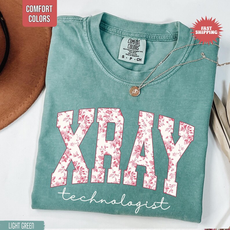 May include: Light green t-shirt with the word "XRAY" in large, floral-patterned letters and the word "technologist" below. A silver necklace with a pendant rests on the shirt. The shirt is labeled "Comfort Colors".