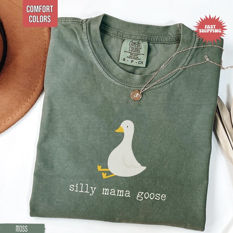 May include: A moss green Comfort Colors t-shirt featuring a white goose illustration with yellow accents. The text "silly mama goose" is printed below the goose. A silver necklace with a pendant is draped across the shirt.