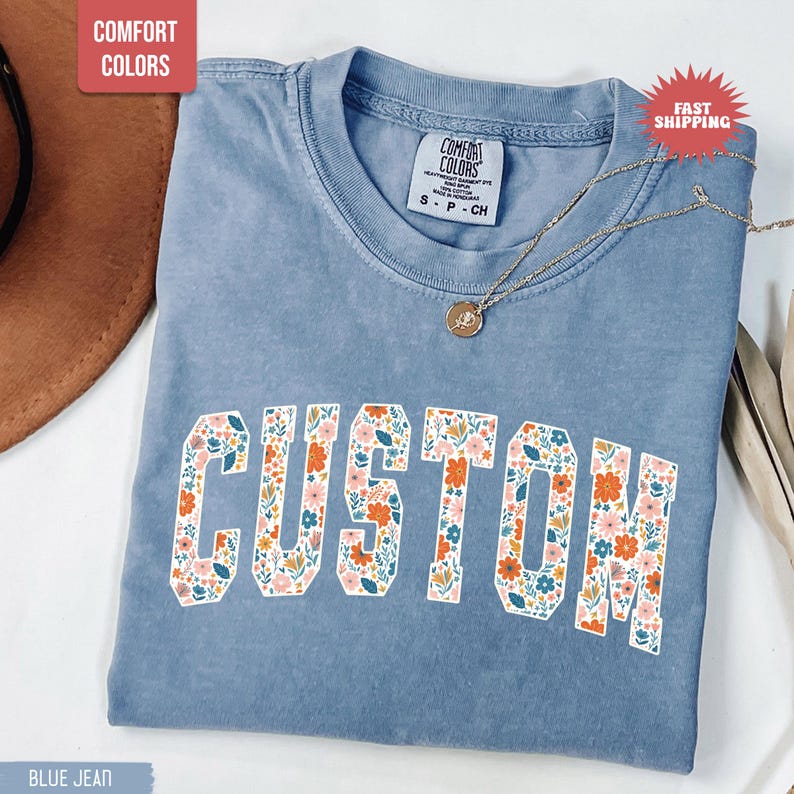 K&ouml;nnte beinhalten: Blaues Jeans-T-Shirt mit dem Wort "CUSTOM" in einem floralen Design. Das Shirt hat einen Rundhalsausschnitt und kurze &Auml;rmel. Eine goldene Halskette mit Anh&auml;nger ist auf dem Shirt drapiert. Das Shirt ist mit "Comfort Colors" gekennzeichnet.
